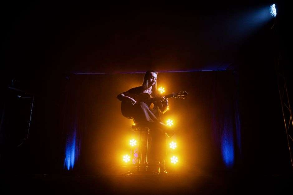 A musician plays acoustic guitar under stage lighting during a live performance