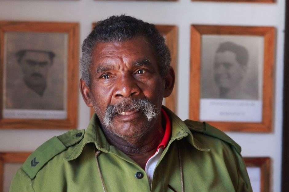 An elderly military veteran sits surrounded by framed photographs from his decades of service