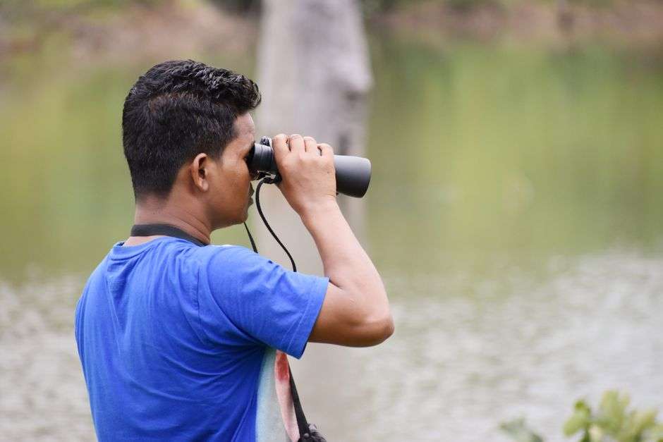 A birdwatcher looks through binoculars near a coastal waterway during a birding outing