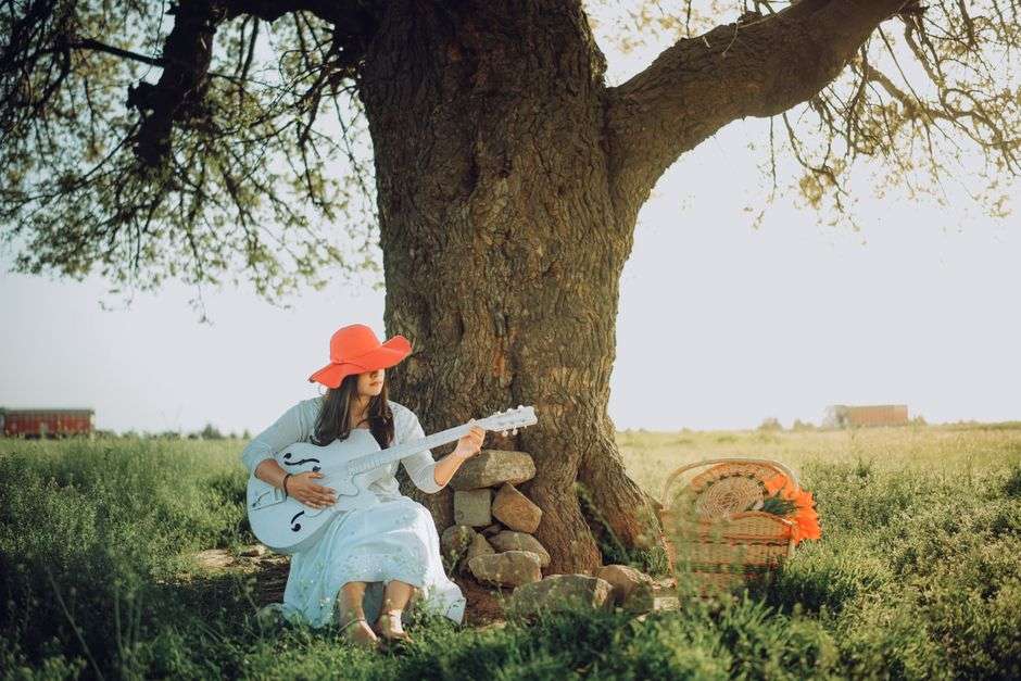 A musician plays acoustic guitar outdoors beneath the shade of a large tree