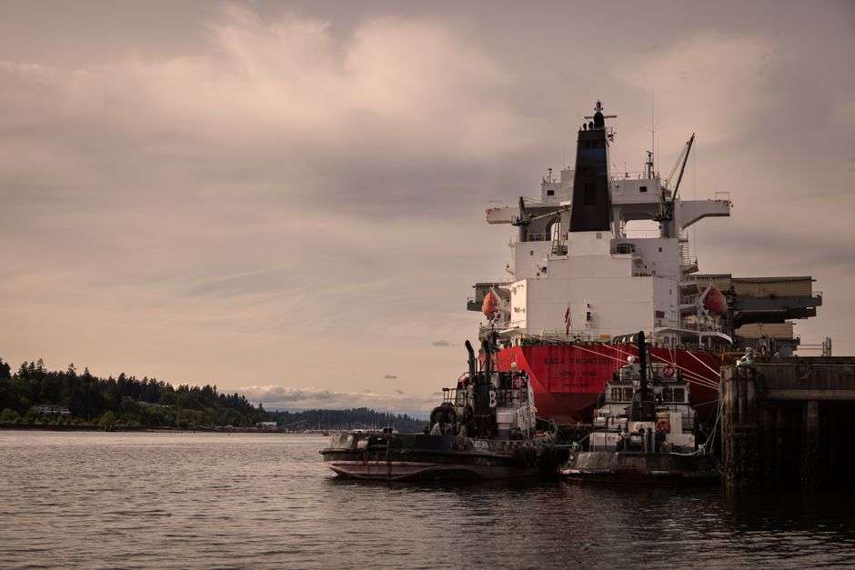 A large ocean liner moored at an industrial river pier, assisted by tugboats