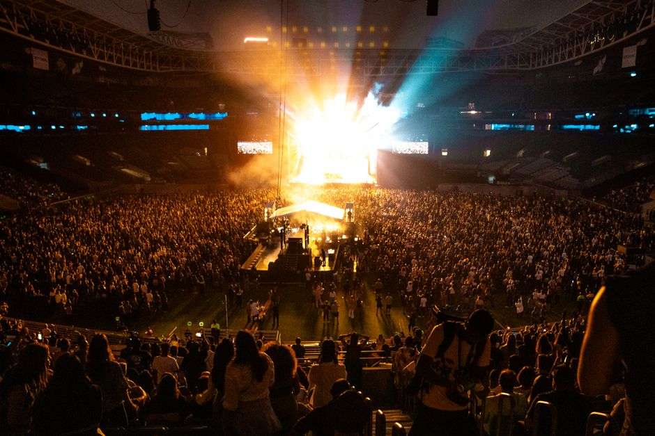 A large crowd gathers at an outdoor amphitheater for a live concert with stage lights