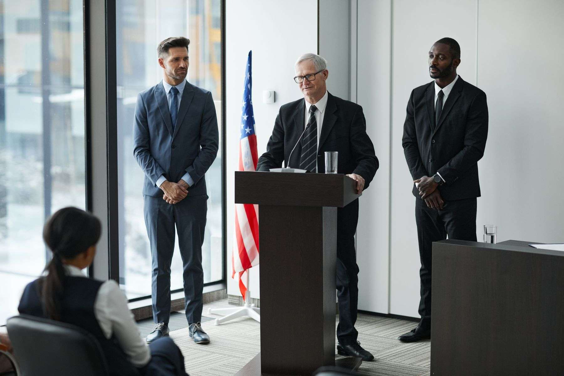 A speaker addresses a room of attendees at a political meeting with an American flag in the background