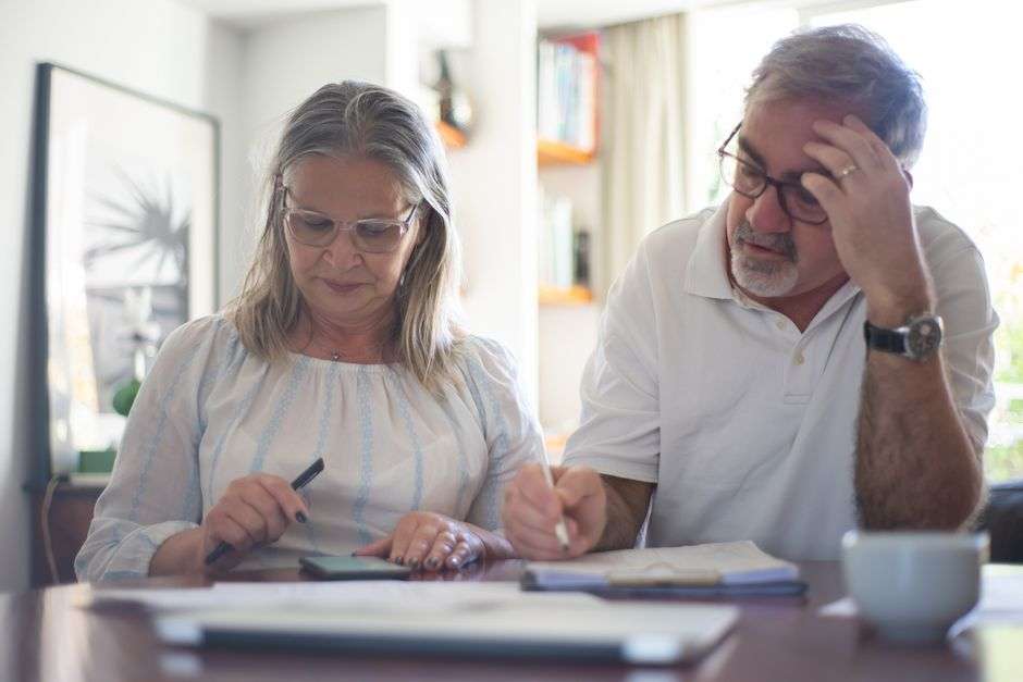 A retired couple sits at a home desk reviewing financial documents and calculating expenses