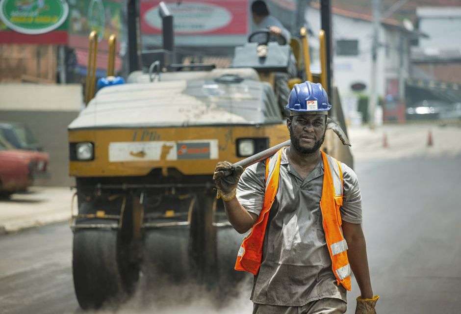 A road construction worker in a safety vest and helmet stands near heavy equipment during a roadwork project