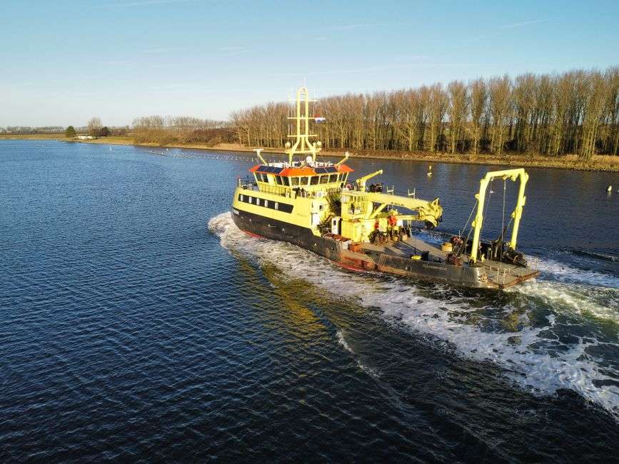 A research vessel equipped with survey gear moves along a calm, tree-lined river