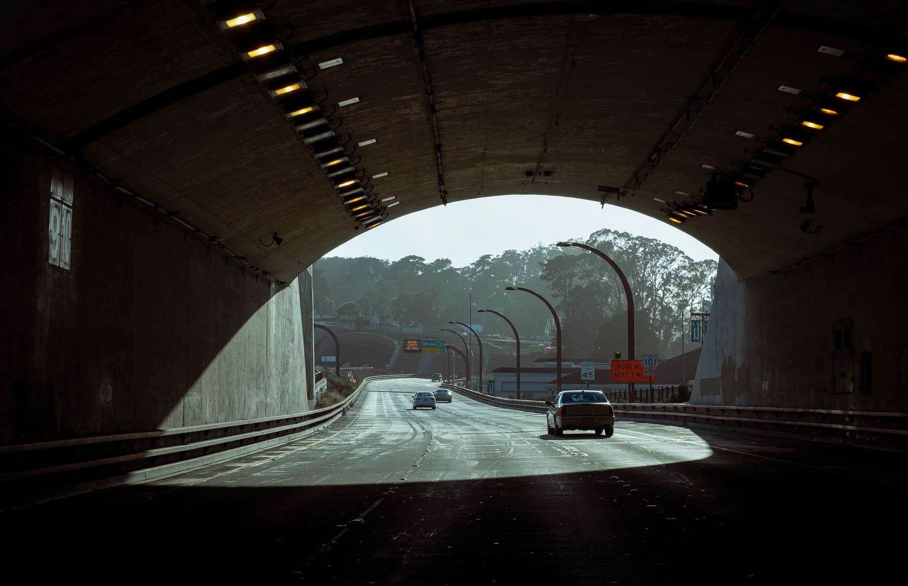 Traffic moving through a dimly lit tunnel with daylight and trees visible ahead