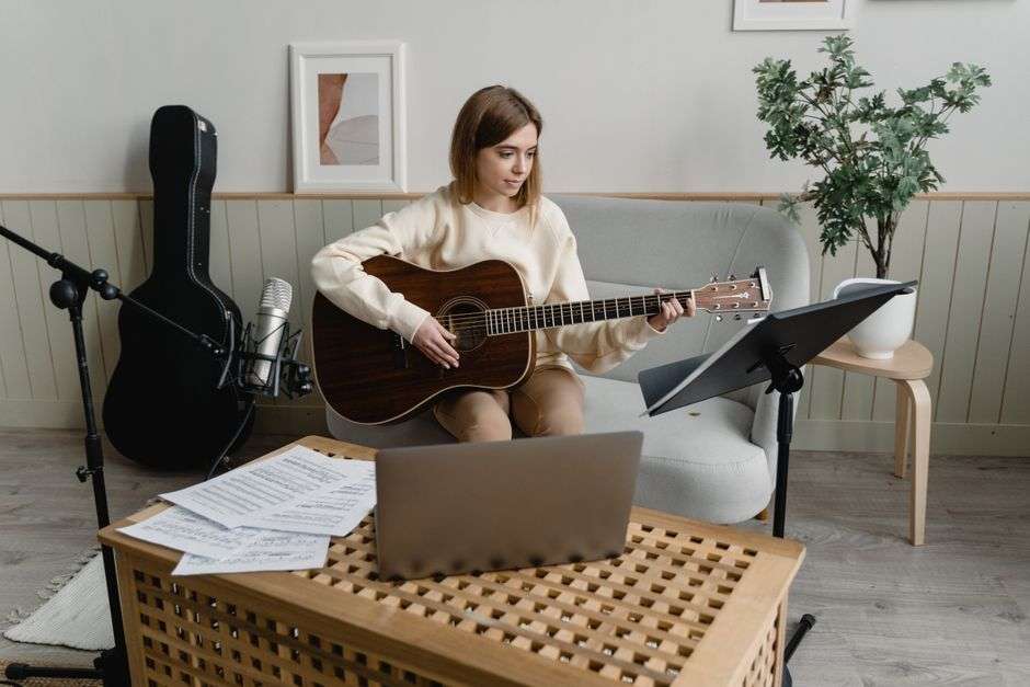 A woman sits at home playing an acoustic guitar and singing into a microphone