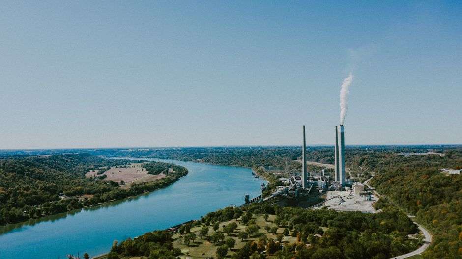 An industrial power plant with tall smokestacks sits along the bank of a wide river