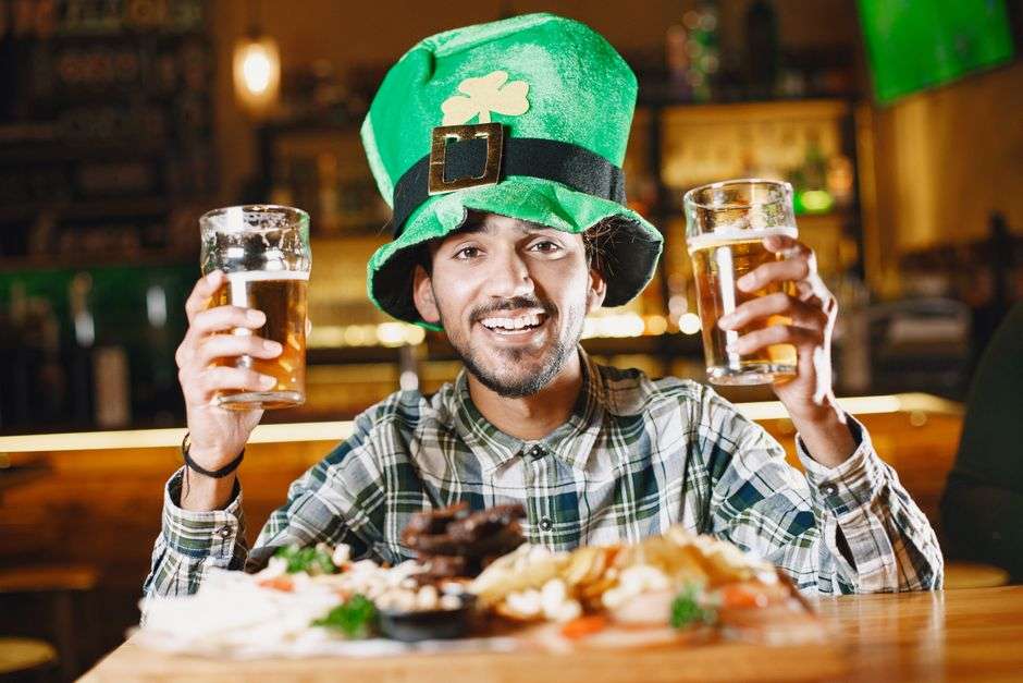 Man wearing a green leprechaun-style hat raises two pints of beer at a pub during a St. Patrick's Day celebration