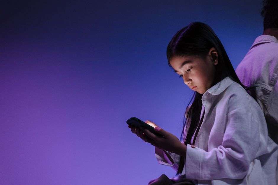 A teenage girl looks at a smartphone screen alone in a dimly lit room