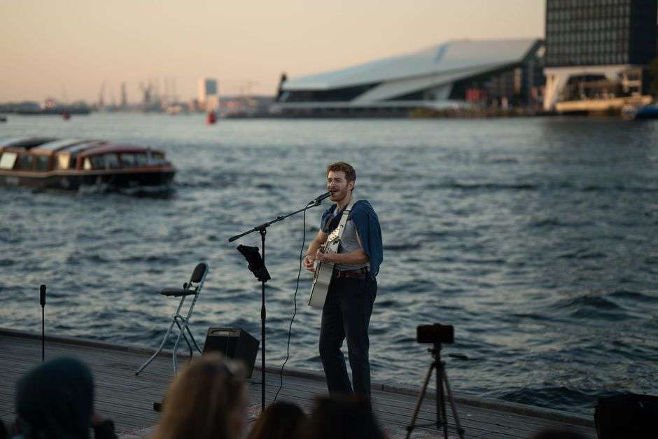A musician performs an acoustic guitar set outdoors near the water at sunset, evoking a laid-back tropical rock atmosphere.