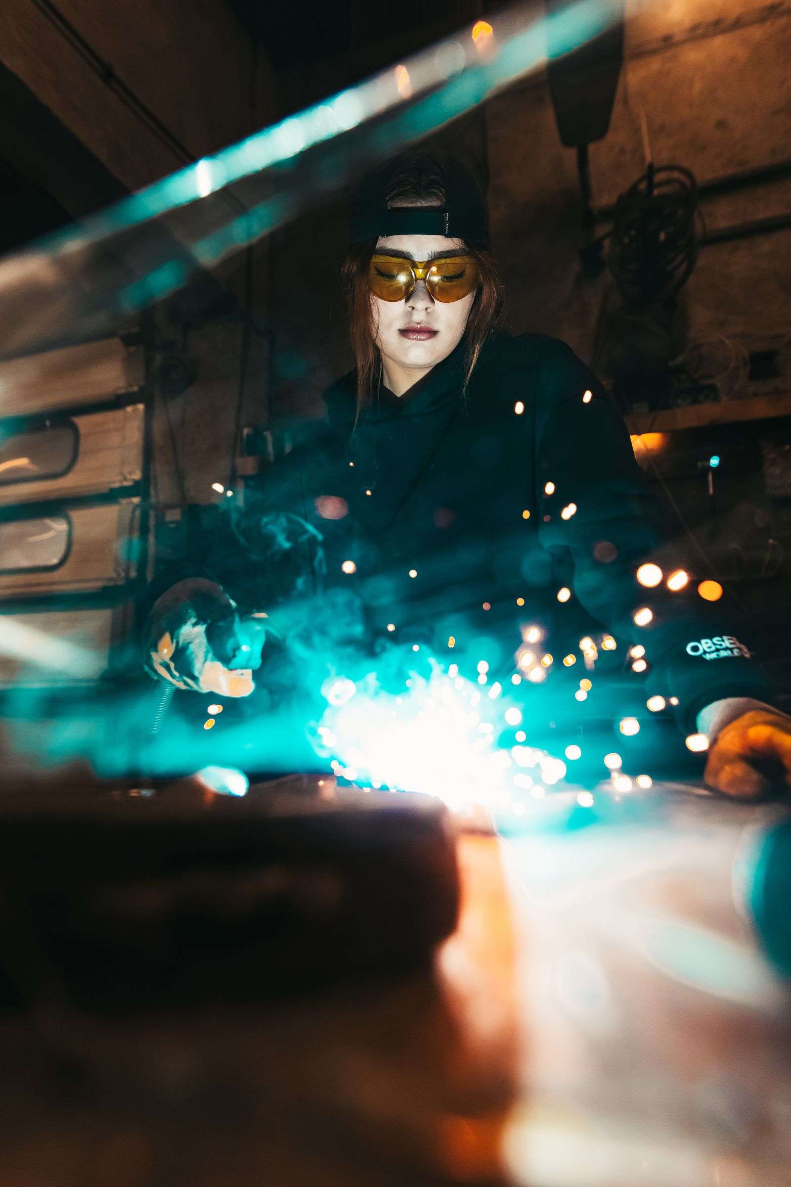 A woman in protective welding gear works with a torch, creating sparks in a training workshop
