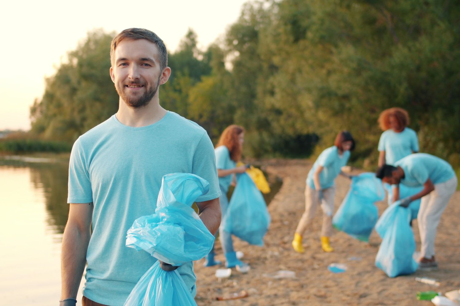 Volunteers in matching t-shirts collect litter in trash bags along a wooded shoreline