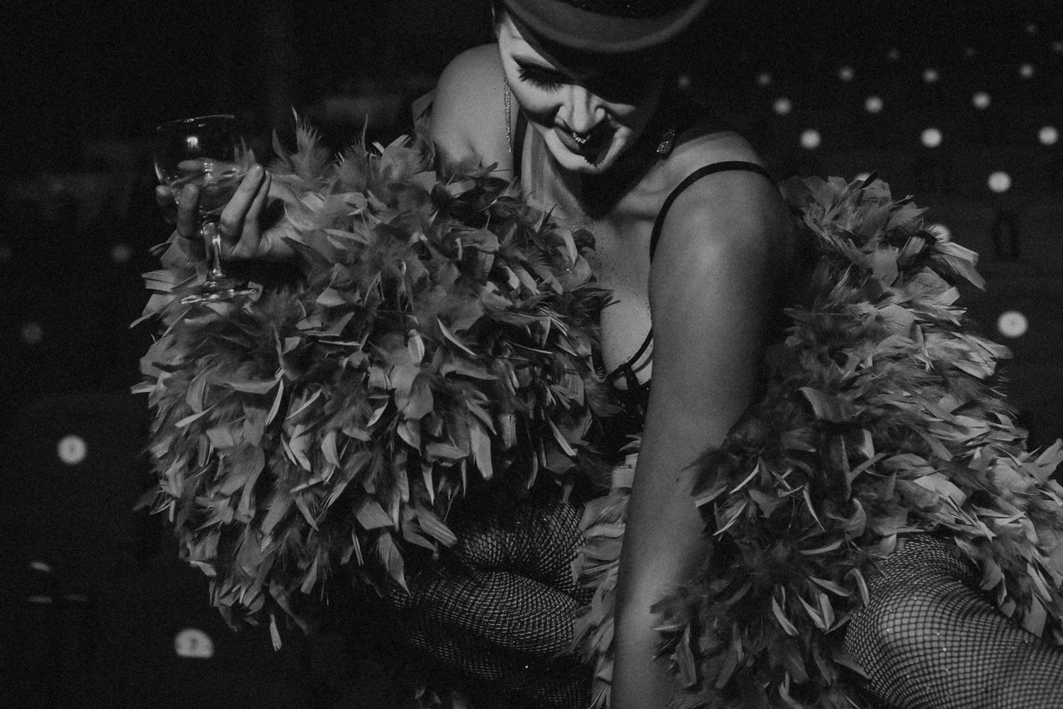 A burlesque performer in a feather boa holds a wine glass under dim stage lighting