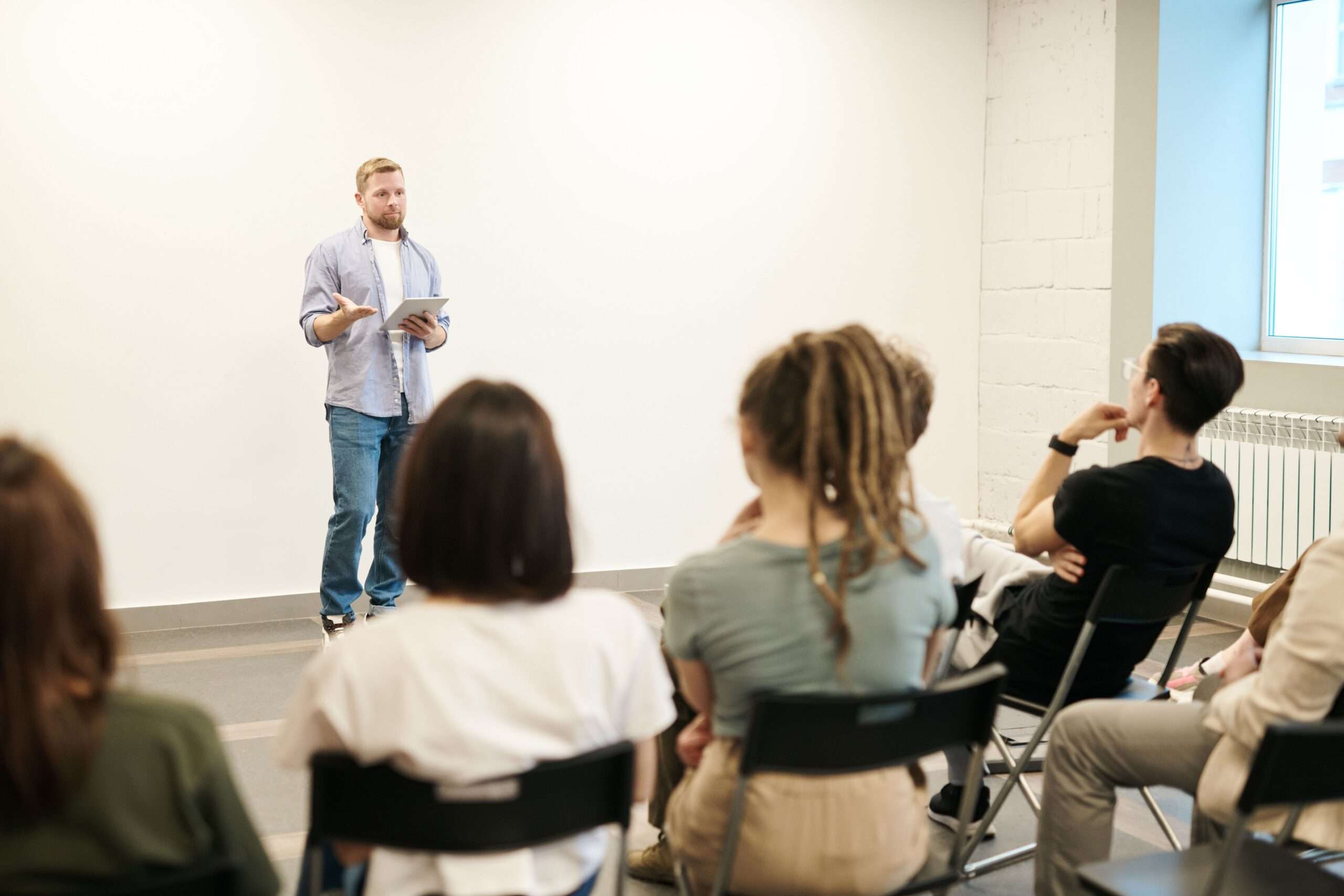A man speaks to a small seated group of adults in a plain indoor room