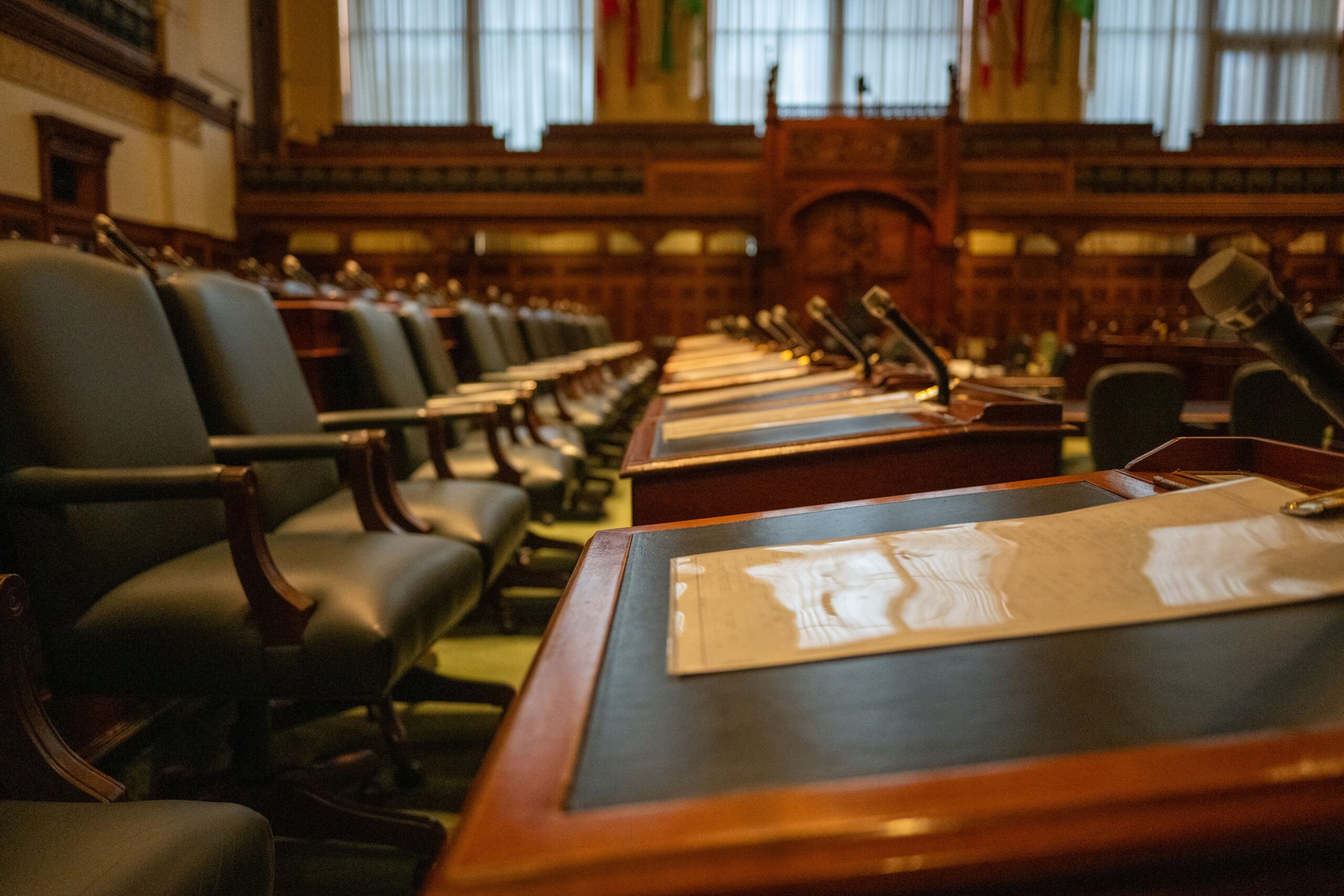 Empty legislative chamber with rows of wooden desks, leather chairs, and microphones