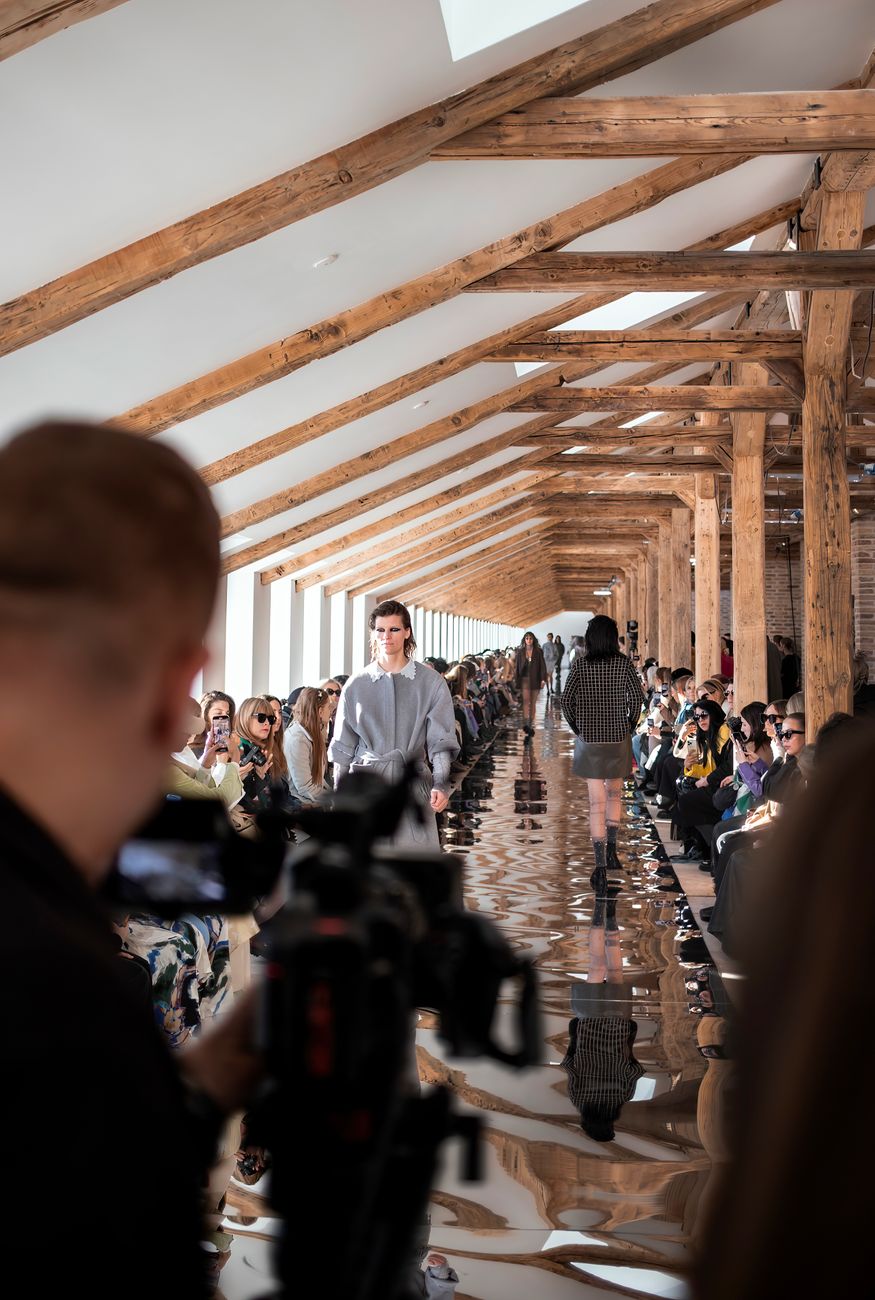 Model walking a runway during a fashion show as an audience watches and photographs the show
