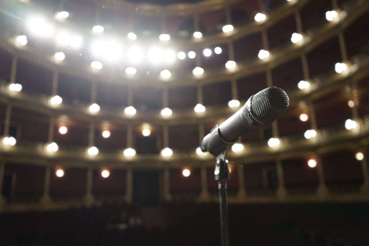 A microphone stands on an empty theater stage facing rows of balcony seating