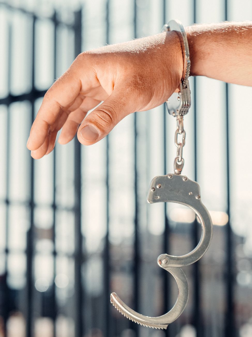 A handcuffed hand and open handcuff dangling in front of metal jail bars