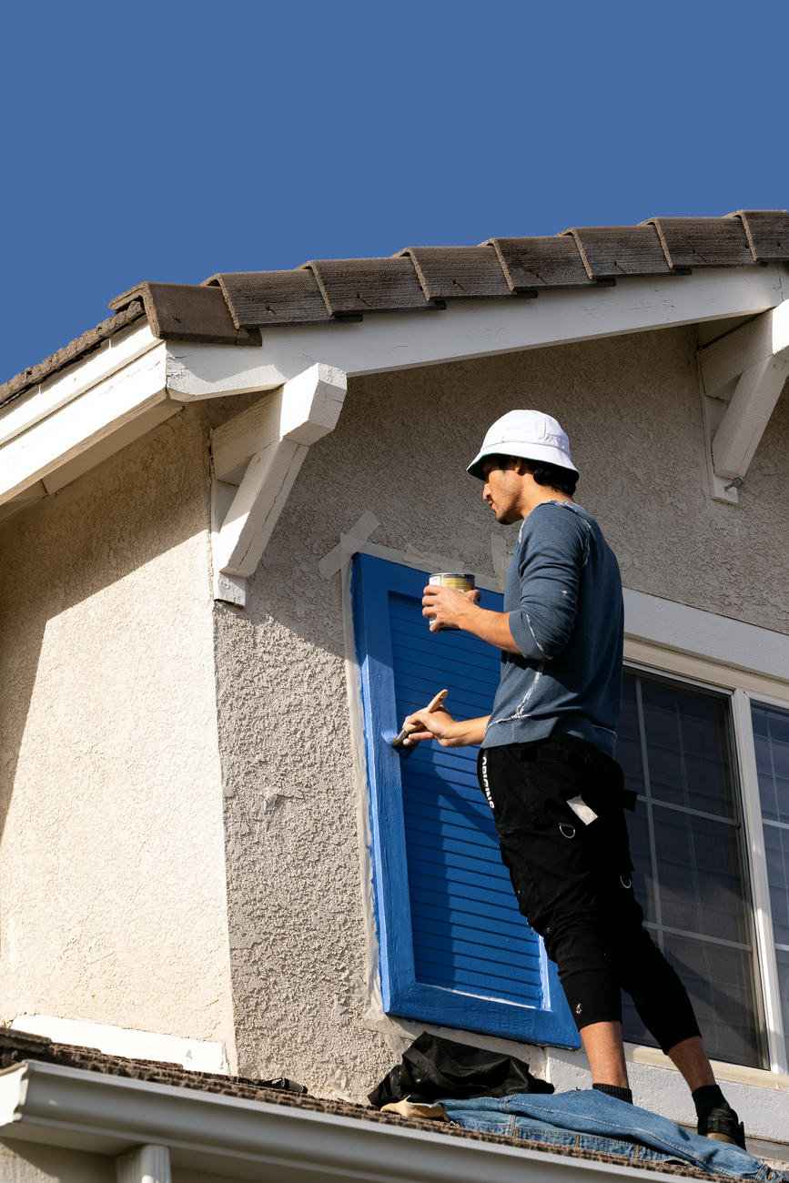 Person on a rooftop painting a house shutter blue during exterior home repairs