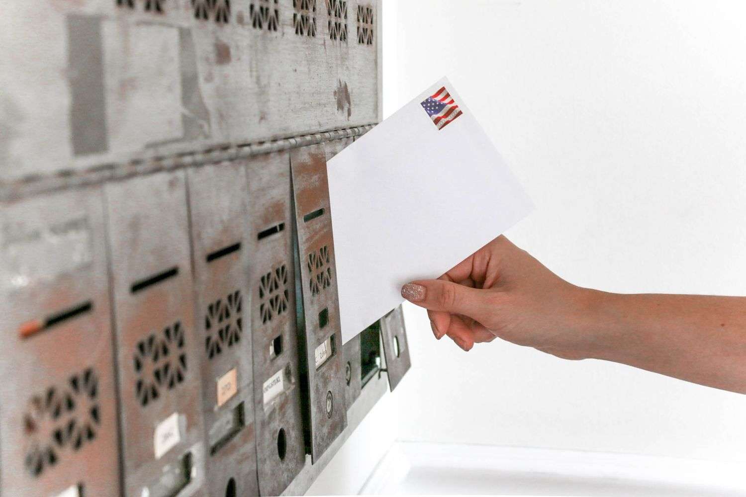 A hand holds a white envelope with a U.S. flag stamp near a wall of metal apartment mailboxes