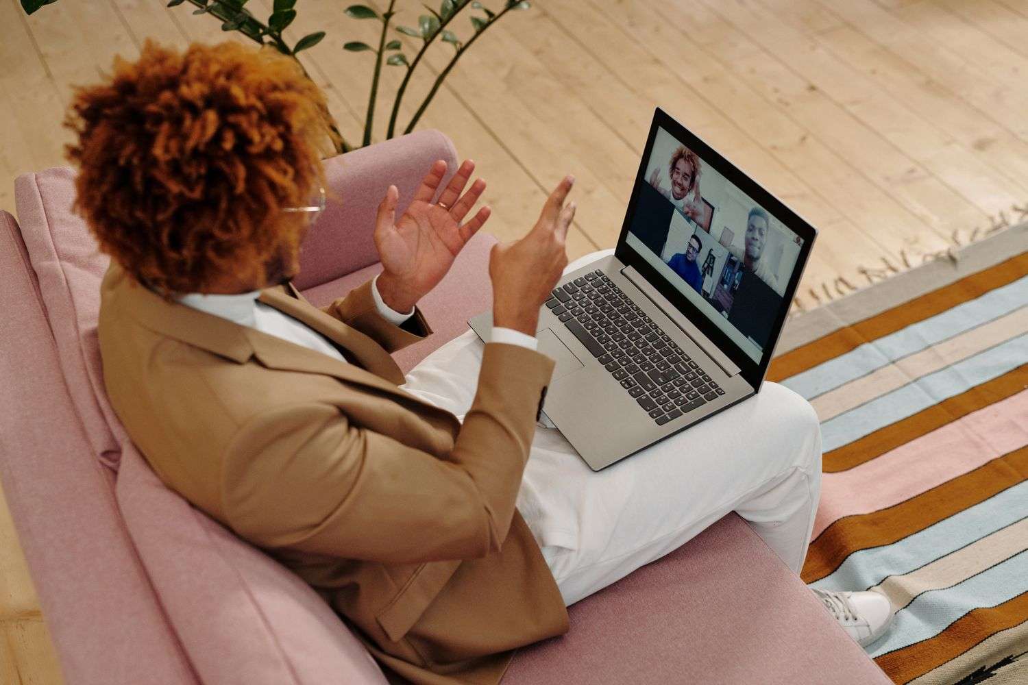 A person sits on a couch participating in a video call on a laptop, with several other participants visible on screen