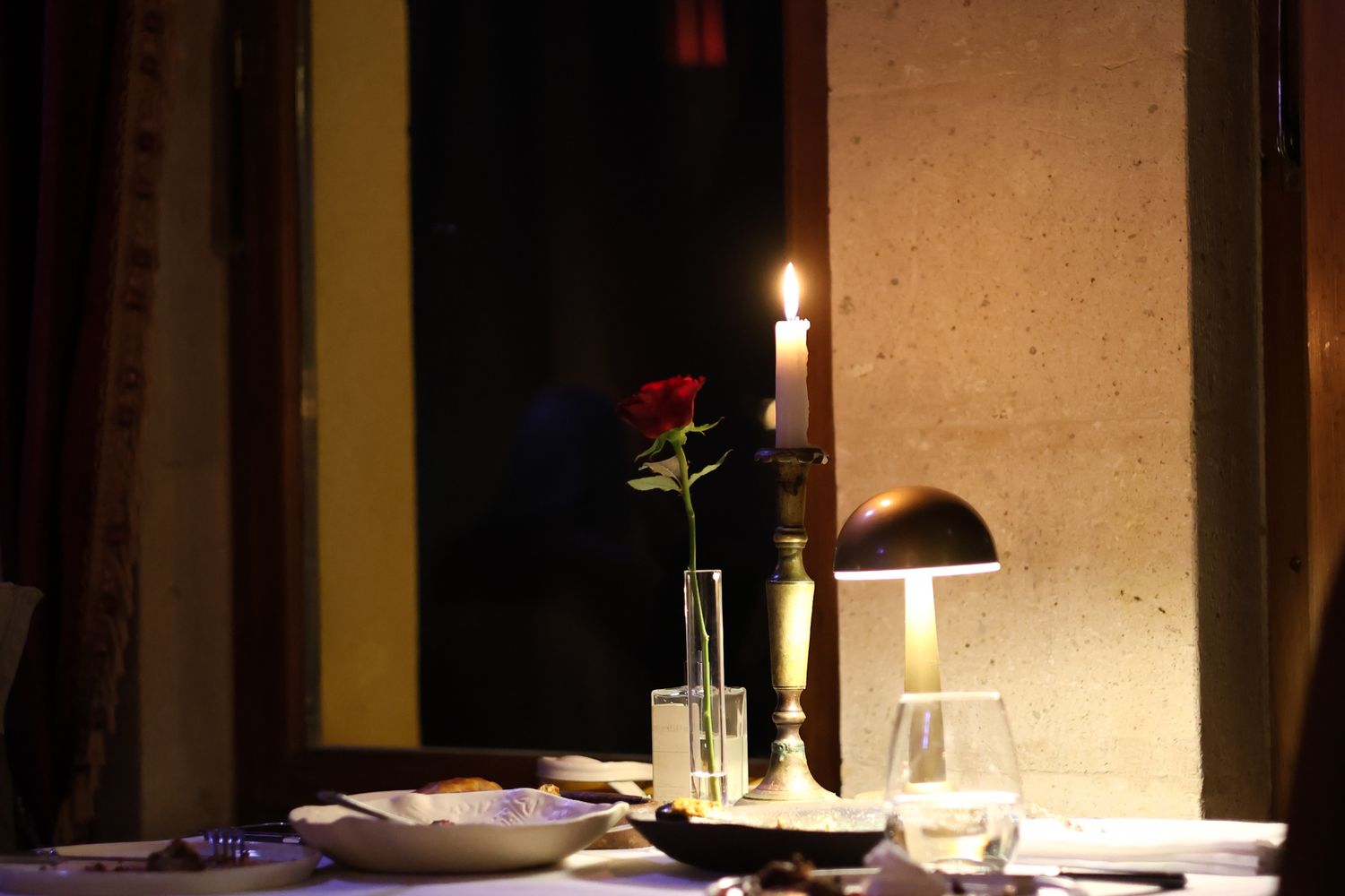 A candlelit restaurant table set with a single red rose in a vase, a lit candle in a brass candlestick, and a glass of wine