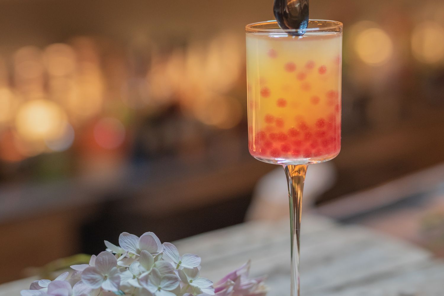 A colorful layered cocktail with red garnish in a stemmed glass, with flowers in the foreground and a dimly lit bar blurred behind