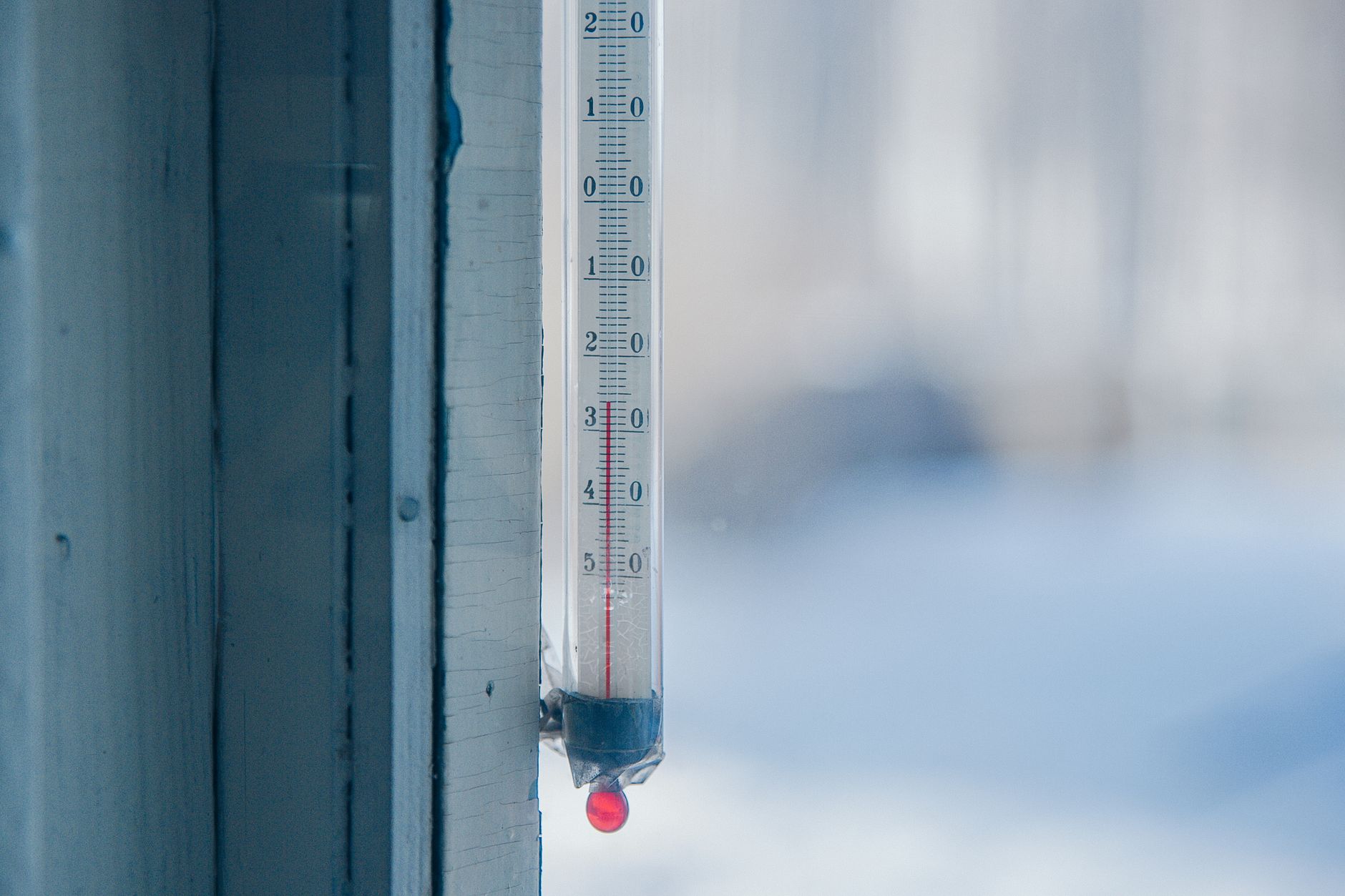 A mercury thermometer mounted on a window frame showing a sub-freezing reading, with a snowy scene blurred in the background.