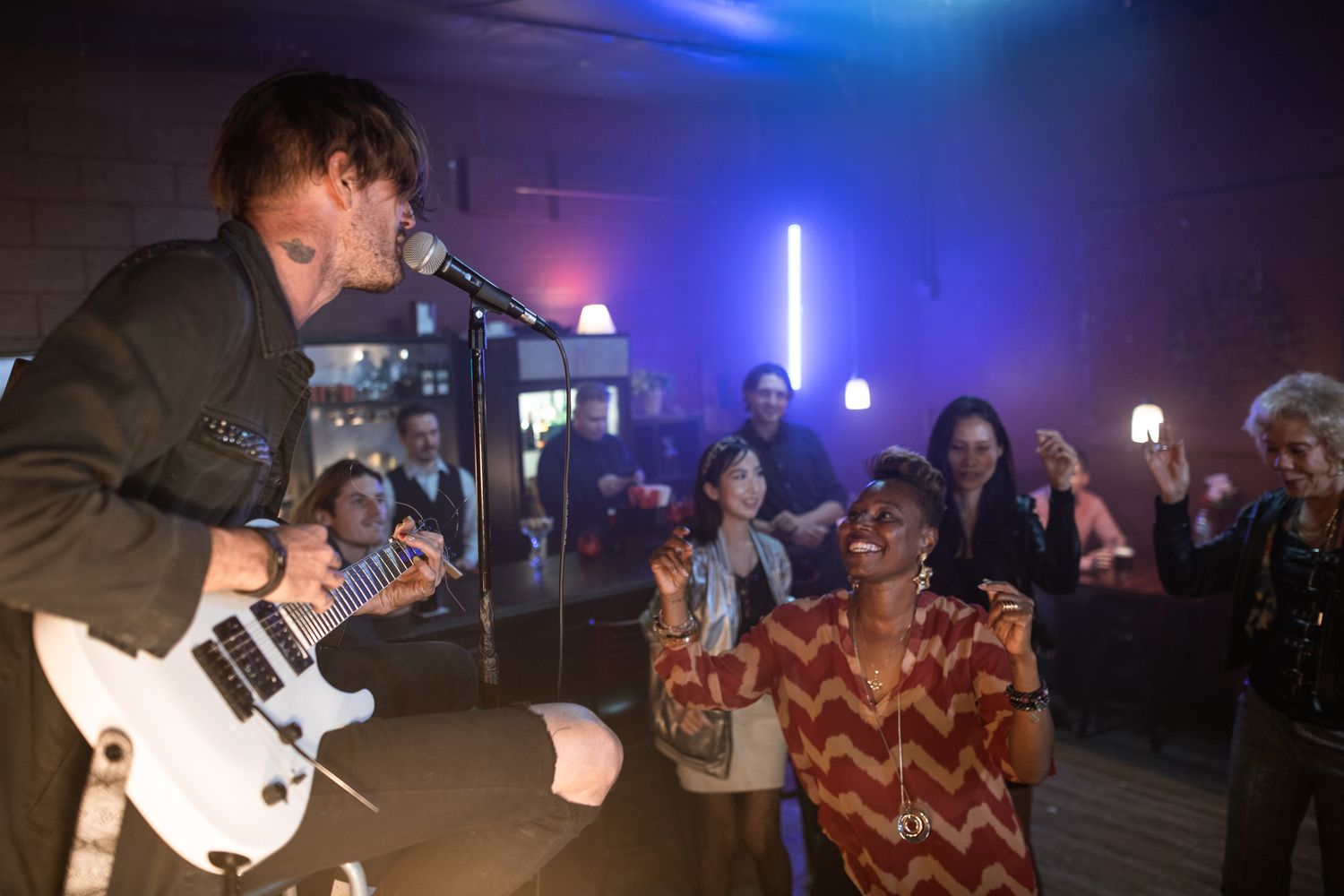 Musician singing and playing guitar on stage while a crowd dances at a bar