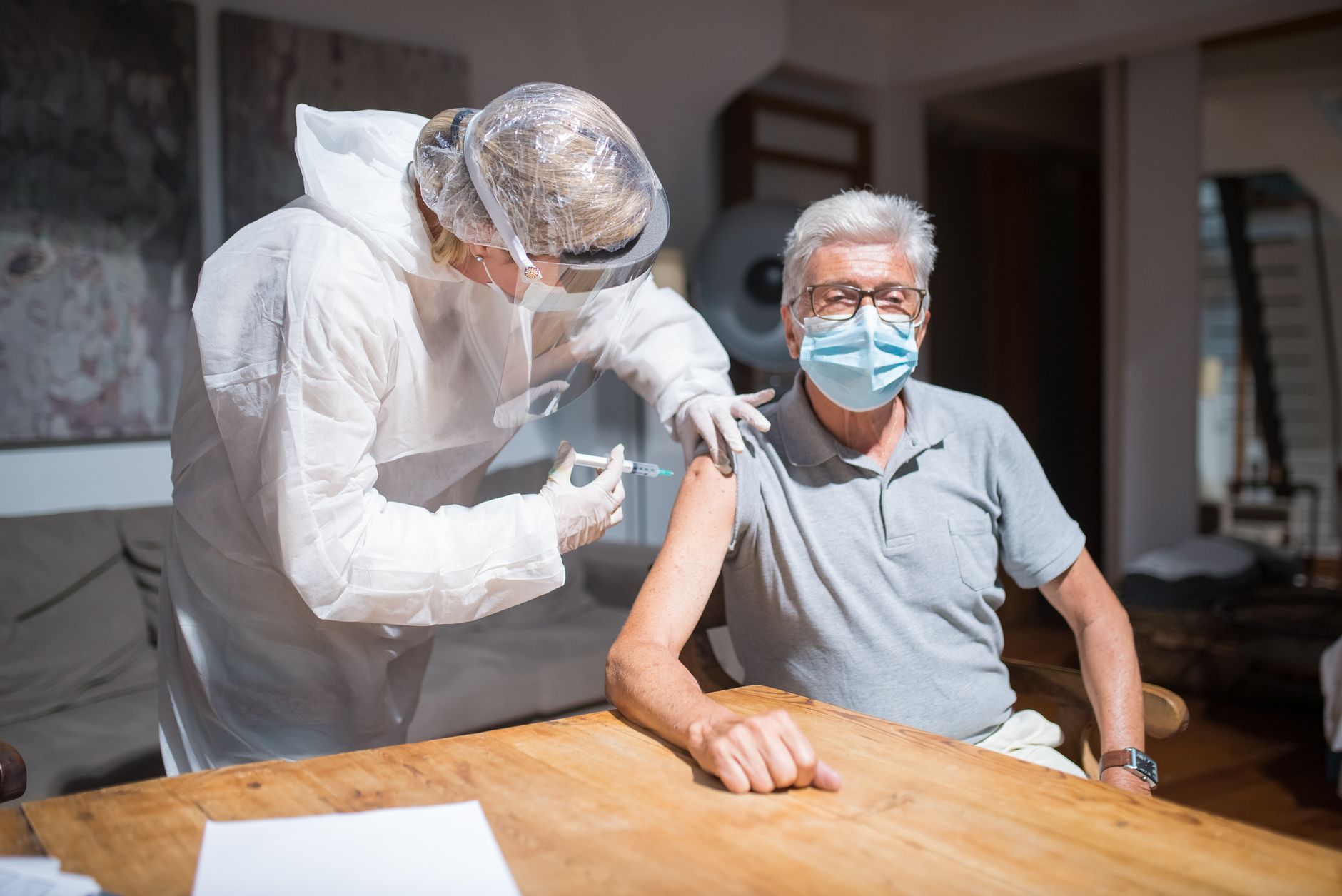 A healthcare worker wearing protective equipment gives a vaccine injection to a seated senior man wearing a face mask.