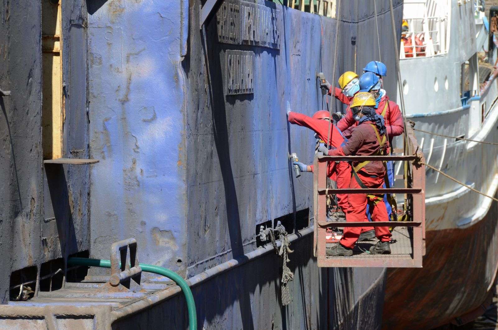 Shipyard workers stand on a hanging platform beside a massive vessel hull
