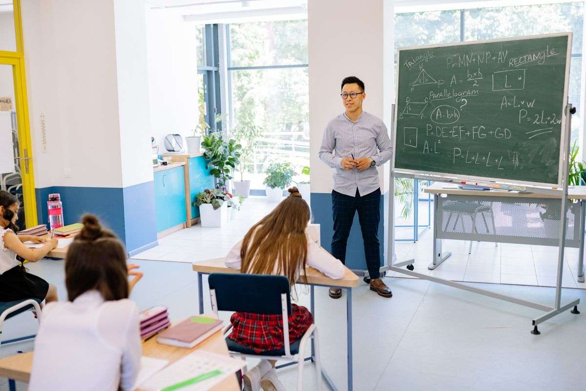 A male teacher standing beside a blackboard in a bright classroom