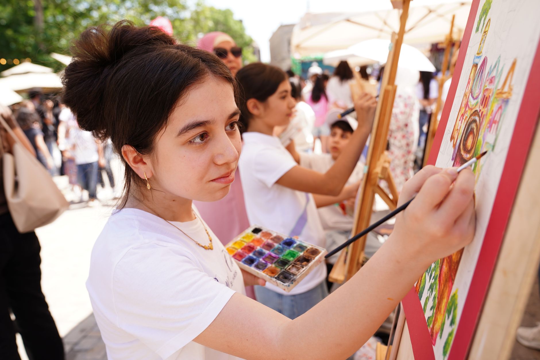 Teenage girl painting on an easel outdoors at an outdoor art event, with another young artist painting in the background