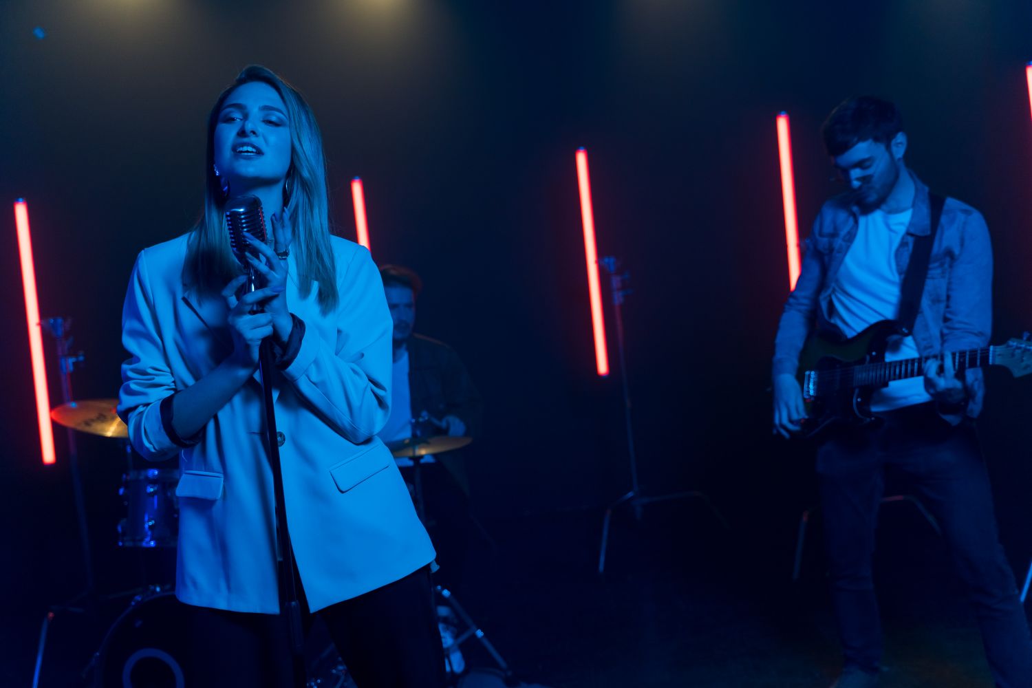 A rock band performs on a dark stage lit with blue and red lighting, with a female vocalist singing into a vintage-style microphone while a guitarist and drummer play behind her.