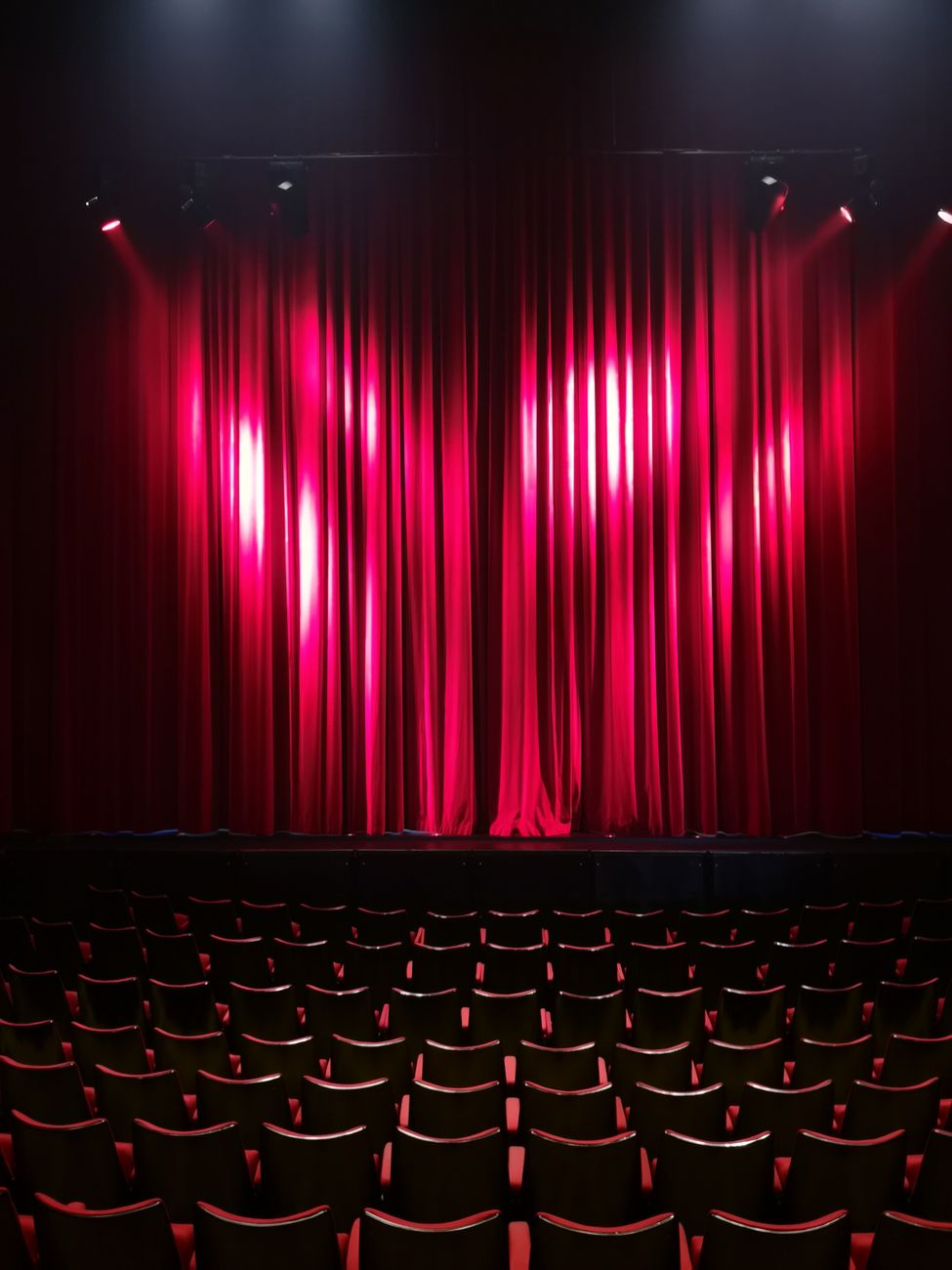 Red stage curtain lit by spotlights above rows of empty theater seats