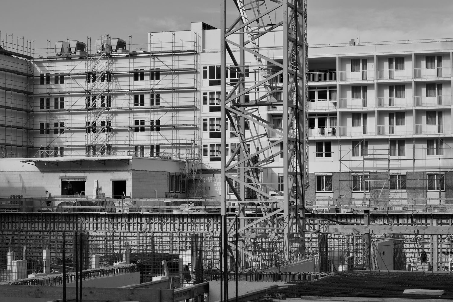 A large multi-story building under construction, with a tower crane, scaffolding, and rebar formwork visible on site