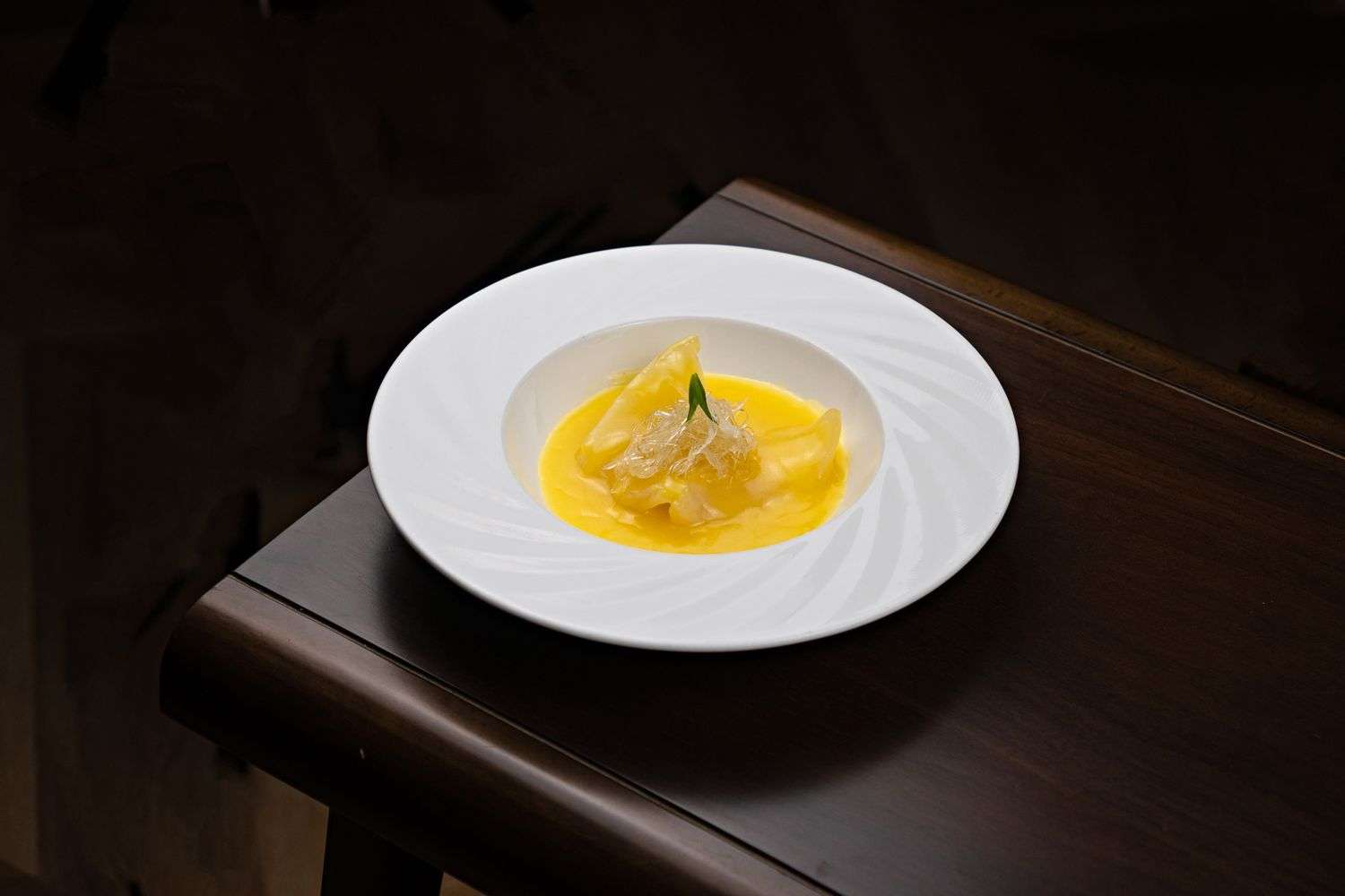 An elegantly plated dish of folded pasta parcels in a yellow cream sauce, garnished with a small green leaf and shredded garnish, on a white patterned plate atop a dark wood table in dim lighting