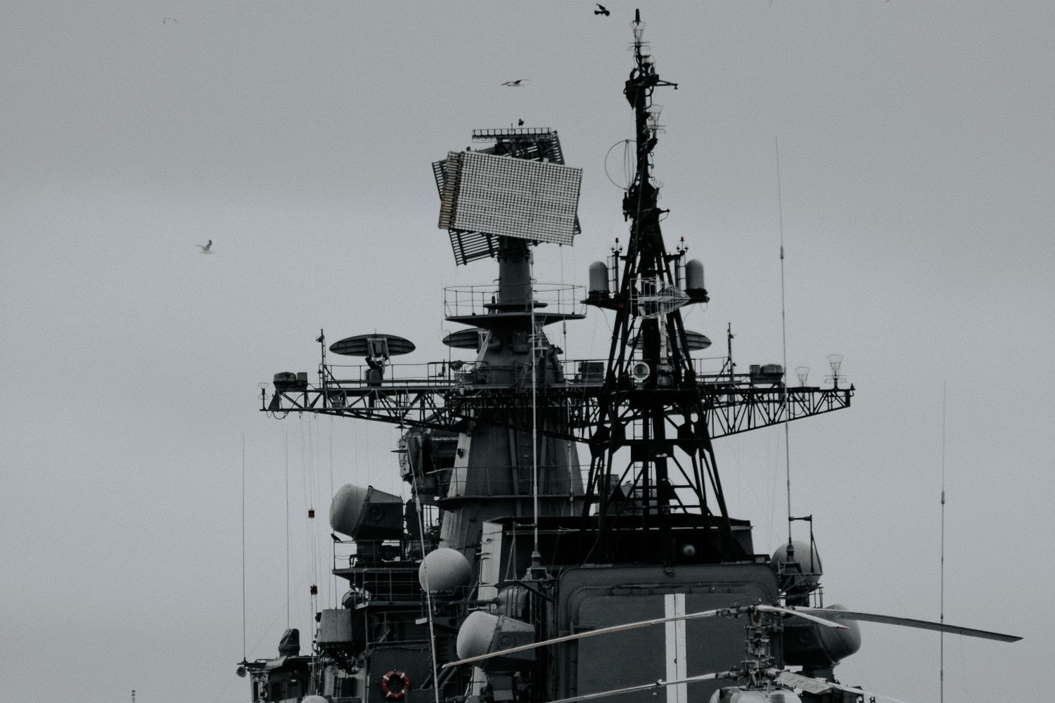 A gray warship's radar mast and superstructure viewed from below against an overcast sky, with seabirds nearby.