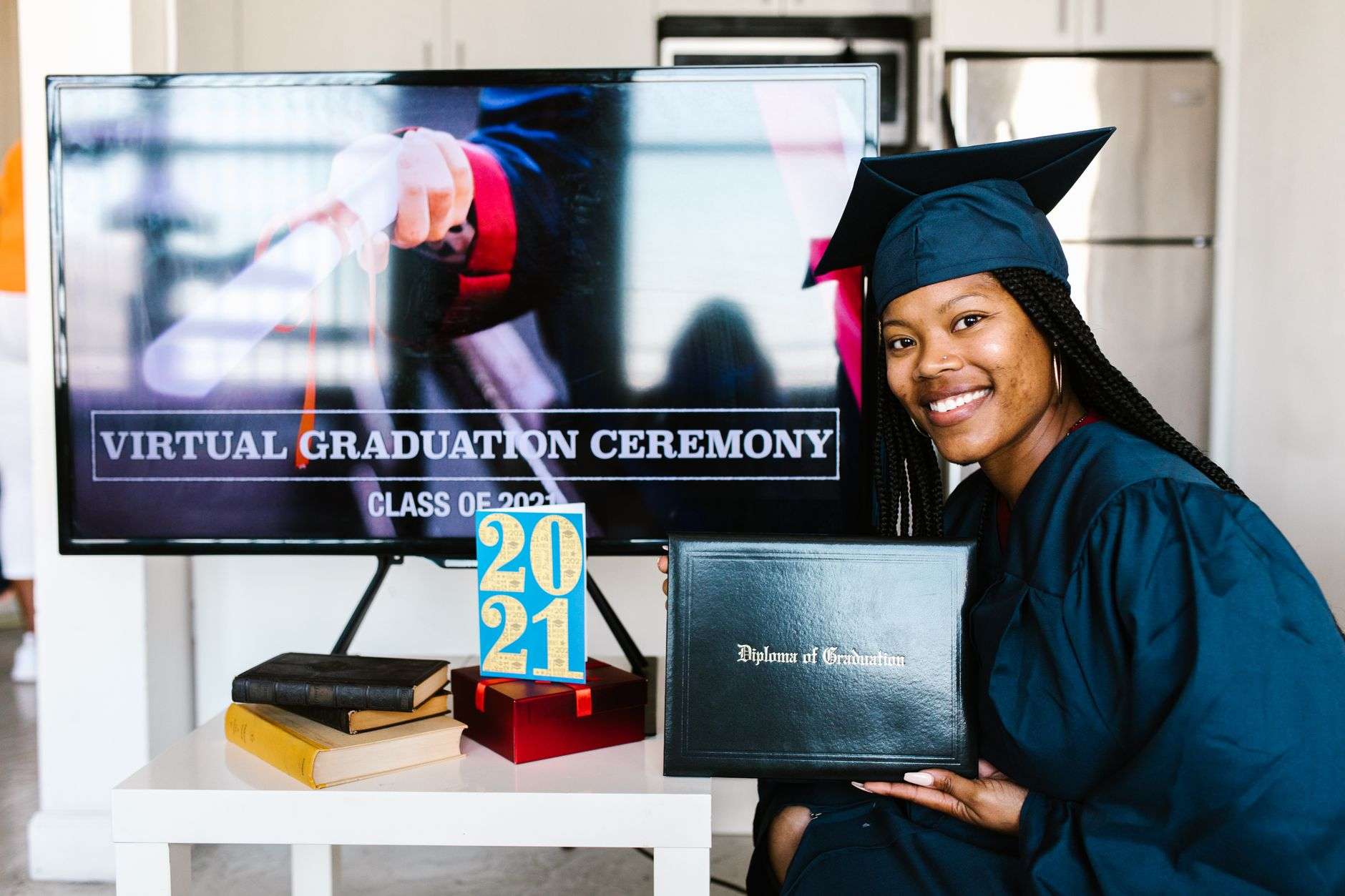 A woman in a graduation cap and gown holds a diploma in front of a television screen displaying a virtual graduation ceremony