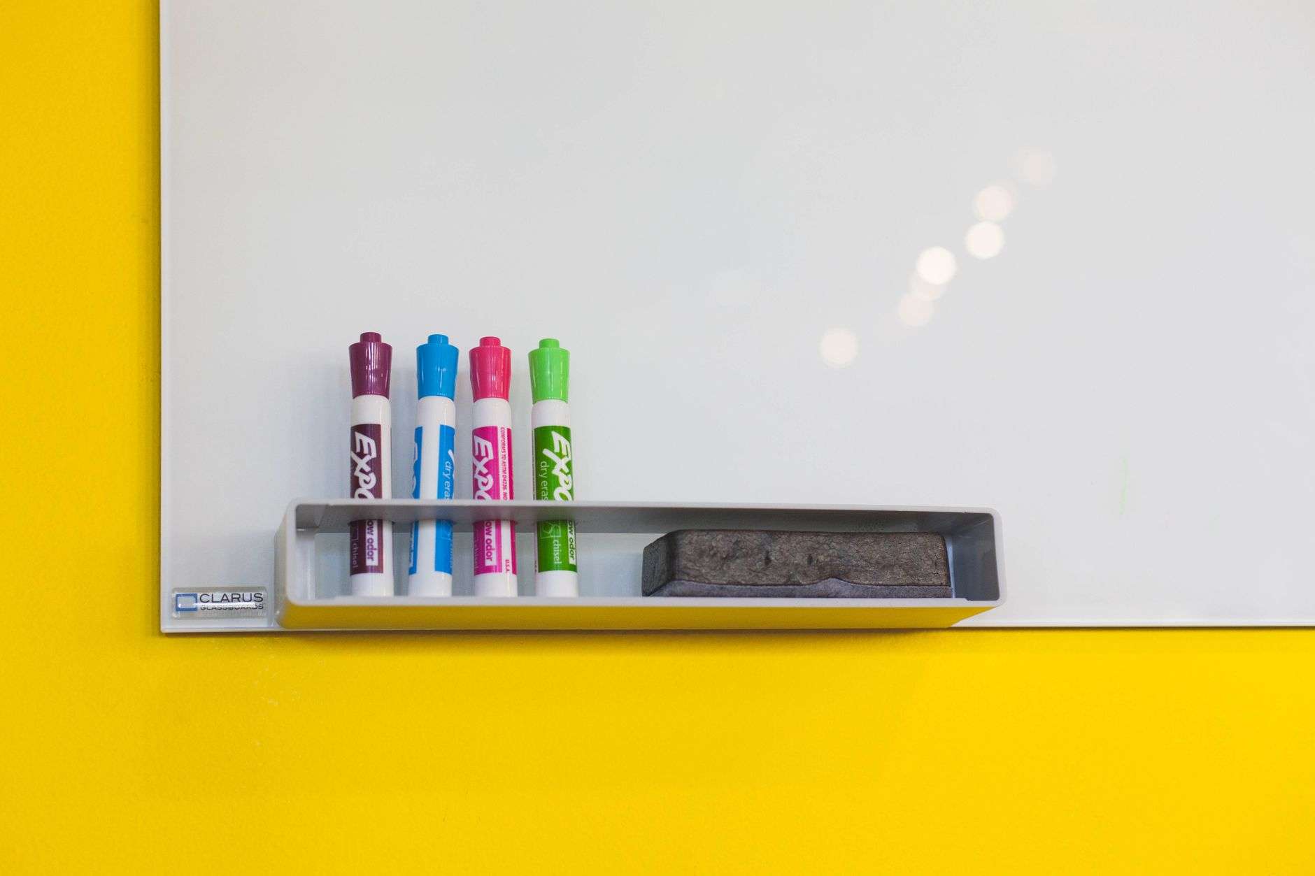 Dry-erase markers and an eraser resting in a whiteboard tray against a yellow wall