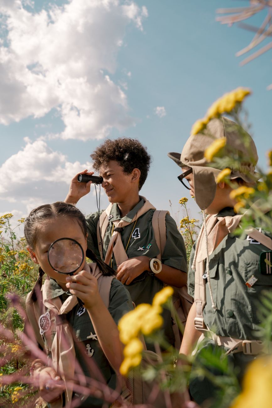 Children in outdoor uniforms examining plants with a magnifying glass and binoculars in a field
