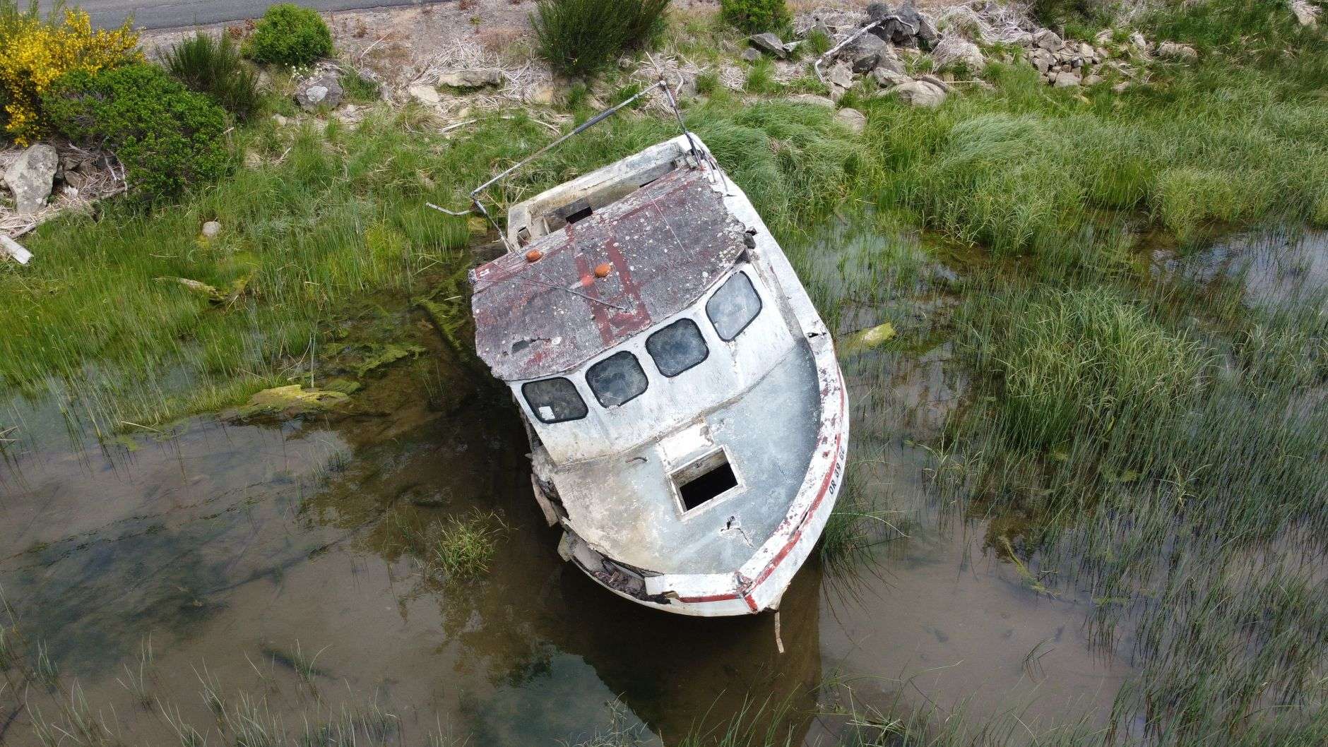 A weathered, partly submerged abandoned boat resting in shallow coastal water near a marsh