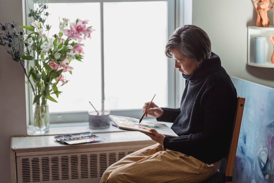 An older woman painting on a canvas at an easel near a window