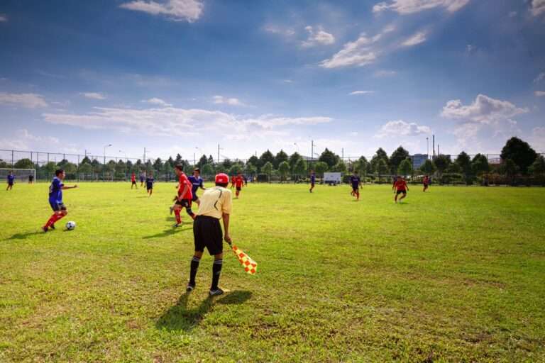 Amateur soccer players in red and blue jerseys chase the ball on a grass field during a daytime match