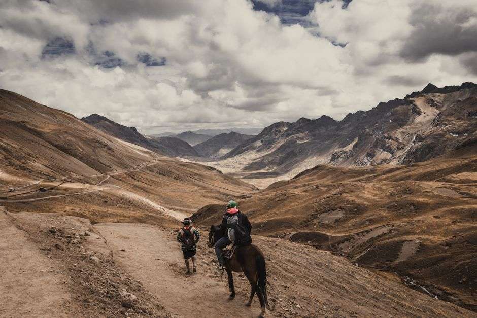 A rider on horseback crossing a mountain trail with peaks in the distance