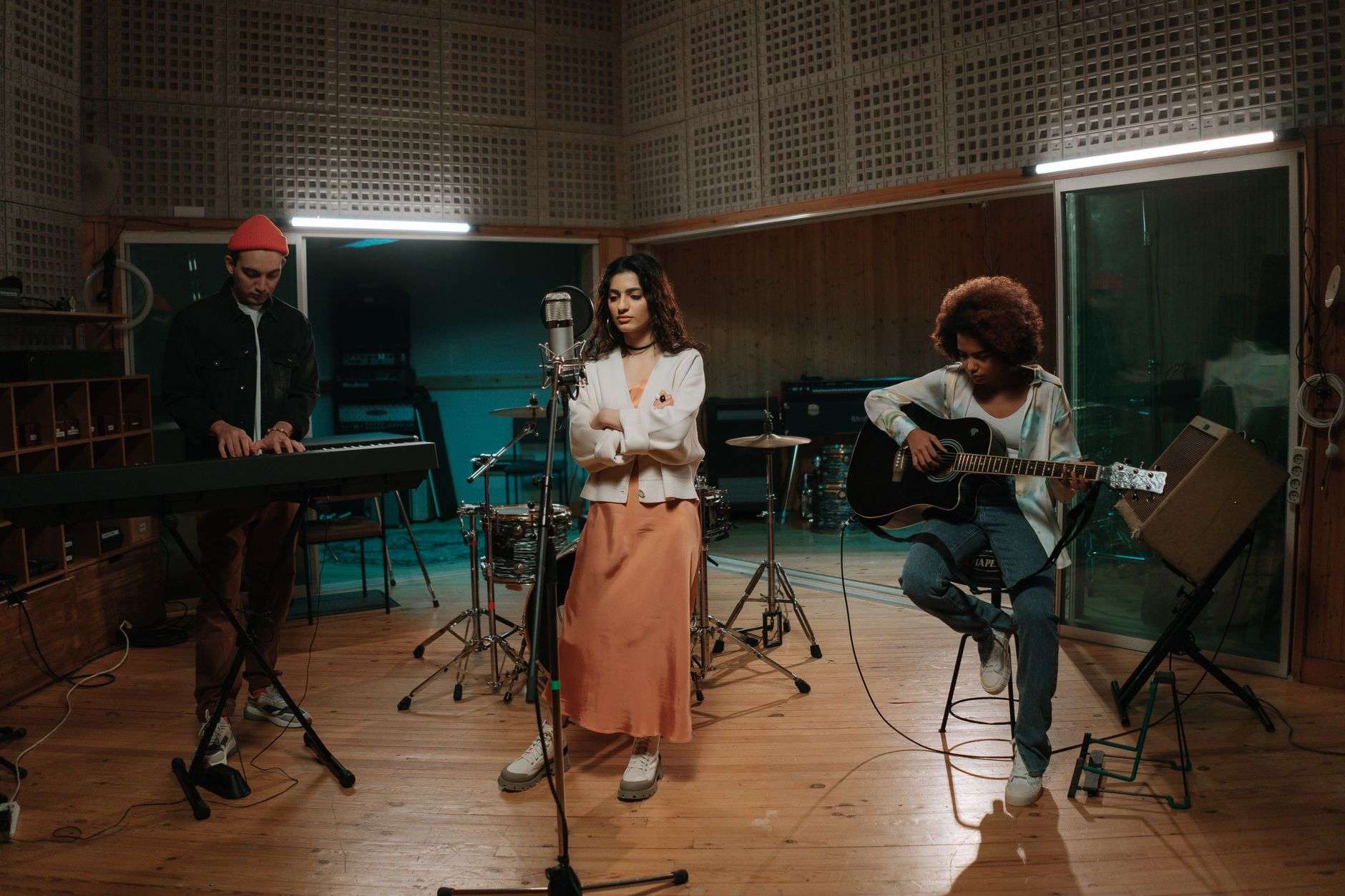 Three musicians performing in a recording studio, with a keyboardist, a vocalist, and a guitarist gathered around microphones and a drum kit