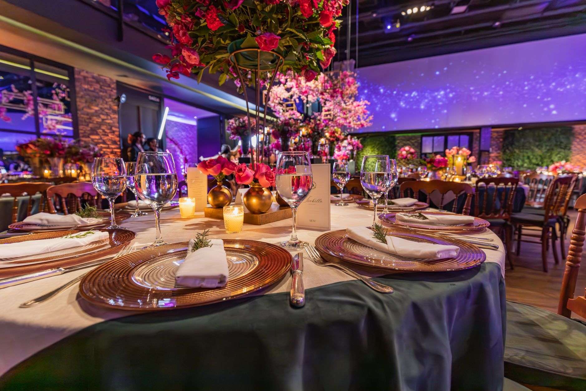 A banquet table set with gold chargers, wine glasses, candles and red floral centerpieces