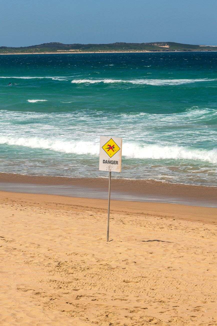 A yellow diamond danger sign with a rip current warning symbol posted in the sand near breaking ocean waves